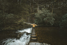 Side View Of Male Tourist In Bright Orange Jacket Walking On Footbridge And Crossing River With Water Flowing Through Stepping Stones In Forest Of Northern Ireland
