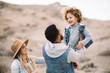 © Gabriel Trujillo/ADDICTIVE STOCK - Cheerful multiracial parents in stylish casual clothes holding smiling adorable curly ethnic toddler and having fun at sandy landscape