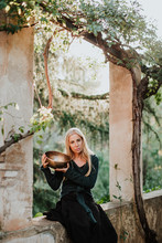 Charming Blond Female In Long Gown Clothes Looking At Camera And Holding Metal Musical Bowl While Sitting In Ruins Of Old Castle In Historic Place