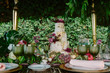 © Gabriel Trujillo/ADDICTIVE STOCK - White and gold tiered cake with flowers placed on wedding table with plate and glasses decorated with fruits and flowers against green trees
