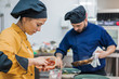 © Javier De La Torre/ADDICTIVE STOCK - Side view of young female cook in yellow uniform holding pot with sauce while working together with male colleague in restaurant kitchen