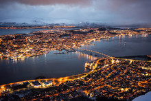 Magnificent Scenery Of City With Golden Lights Located On Island And Shores Of Strait Against Foggy Hills Covered With Snow Under Lush Clouds In Winter Night