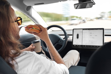 Young adult man sitting in car, eating fast food, drinking beverage