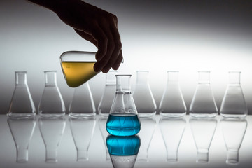 Hand pouring yellow in to blue liquid in scientific laboratory glass erlenmeyer flask with glassware equipment on reflective surface.