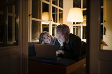 Happy Senior Couple Lying On Couch At Home At Night Using Laptop