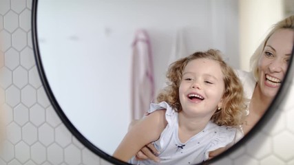 Wall Mural - A cute small girl with mother in bathroom indoors at home, looking in mirror.