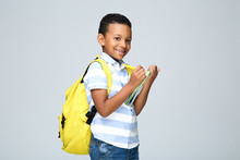 Young African American School Boy Writing In Notepad With Backpack On Grey Background