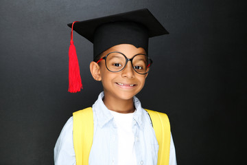 Wall Mural - Young African American school boy in graduation cap with backpack on black background