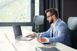 © gorynvd - Successful bearded businessman in glasses and in blue suit works with laptop in modern office.