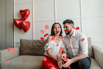 Wall Mural - happy couple man and woman in love with heart shaped confetti in the air sitting on couch at home