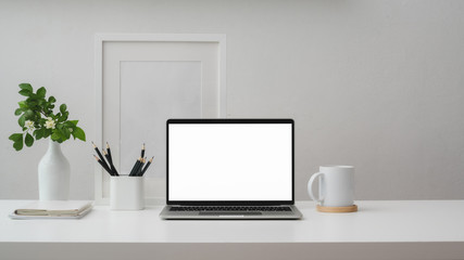 Cropped shot of workspace with blank screen laptop, frame, pencils, coffee cup and vase on white table with white wall