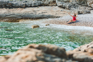 Wall Mural - young pretty woman in red swimsuit sitting at rocky beach with phone taking selfie picture