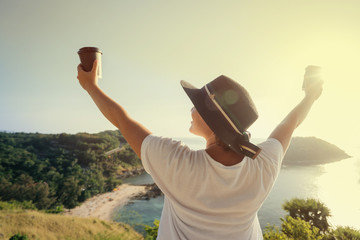 A young woman in a hat holds two disposable paper cups with coffee on the background of a beautiful tropical landscape of mountains and the sea.