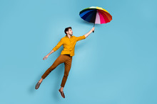 Full Length Photo Of Crazy Guy Jumping High Holding Colored Bright Umbrella Flying Up Higher Wear Yellow Shirt Trousers Isolated Blue Color Background
