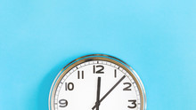 Top Of Big Plain Wall Clock On Pastel Blue Background. Five Past Twelve O'clock. Top View Copy Space, Time Management Or School Concept And Summer Or Winter Time Change, Opening Hours, Lunch Break