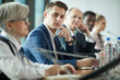 © AnnaStills - Young businessman in suit sitting at the table and listening to his colleagues during business conference