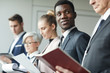 © AnnaStills - Young African businessman in formalwear holding report and standing in a row with other business people at office