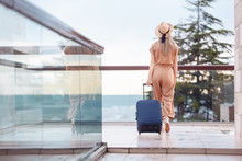 Smiling Young Stylish Beautiful Woman In Silk Overalls And Hat With Suitcase Check In Hotel Room With Terrace Overlooking Sea And Nature. Tropical Vacation Concept. Expensive Luxury Lifestyle, Trip
