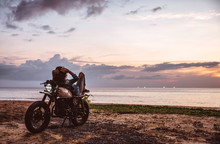 Beautiful Girl Having Fun Driving Her Custom Cafe Racer Motorcycle, Enjoying The Sunset On The Beach