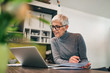 © bnenin - Doing paperwork at home. Senior woman looking at laptop and holding paper document, portrait.