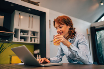 Poster - Side view of a businesswoman drinking juice and using laptop in the kitchen, portrait.