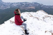 Beautiful European Girl In A Red Park, Warm Jacket, Jeans, Black Leather Boots And Sunglasses Sits On Top Of A Snowy Mountain And Looks Into The Distance