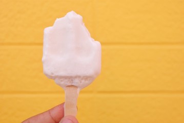 A male hand holding a melting coconut milk ice cream on wooden stick with yellow wall background