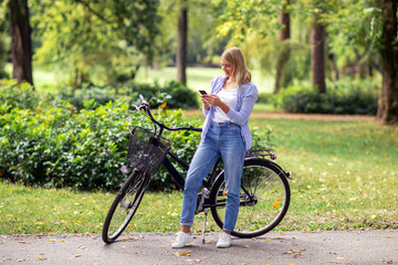 Wall Mural - Beautiful young blonde woman send sms message on mobile phone while sitting on bicycle in the park