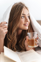 Sticker - Photo of young smiling woman reading book and drinking tea while lying