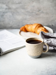 Delicious breakfast - the cup of coffee, croissant and open magazine on the table at the grey background.