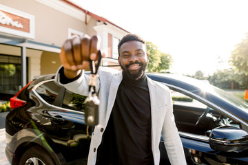 Portrait of African man car seller holding car keys. Attractive cheerful young African man smiling showing car keys to his new auto posing outdoors at the dealership salon