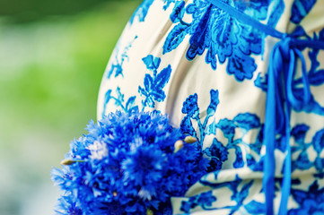 Cropped picture of pregnant woman belly in white and blue dress with bouquet of knapweeds outside in summer time. Expecting baby in summer. Beautiful happy pregnancy and motherhood.