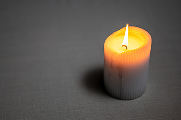 Light beige candle burning with a bright yellow flame. Wax flows down to the table. A small shadow falls on the light table from the candle. Horizontal close-up