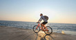© serejkakovalev - Young handsome male in casual wear ride on the colorful bicycle on the morning beach against beautiful sunset and the sea