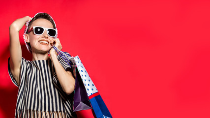 Wall Mural - Happy shopping woman with shopping bags over bright red background wearing hat and sunglasses