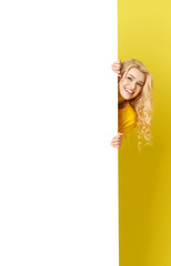 Wall Mural - Young happy woman peeks out from behind a white banner on a yellow background. Point to an empty blank on a form, a copy space for text. vertical shot