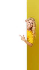 Wall Mural - Young happy woman peeks out from behind a white banner on a yellow background. Point to an empty blank on a form, a copy space for text. vertical shot