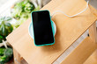 Smartphone in mint silicone case is charged from a wireless charger. The mobile phone is charged on a wooden nightstand or shelf.
