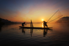 Silhouette Of Asian Fisherman On Wooden Boat In Action Casting A Net For Catching Freshwater Fish In Nature River In The Early Morning Before Sunrise