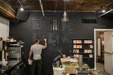 Barista Writing Menu On Coffee Shop Blackboard Wall
