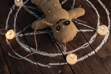 Voodoo doll on a wooden background with dramatic lighting and candles. The concept of witchcraft and black art and the occult. Burlap doll on the background of a drawn star. Stuck pins in the doll.