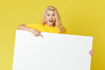 Wall Mural - Young happy woman peeks out from behind a white banner on a yellow background. Point to an empty blank on a form, a copy space for text. Horizontal shot