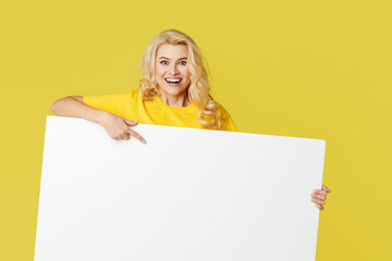 Wall Mural - Young happy woman peeks out from behind a white banner on a yellow background. Point to an empty blank on a form, a copy space for text. Horizontal shot