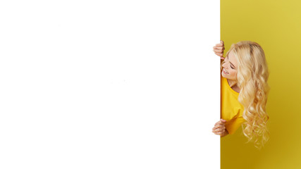 Wall Mural - Young happy woman peeks out from behind a white banner on a yellow background. Point to an empty blank on a form, a copy space for text. Horizontal shot