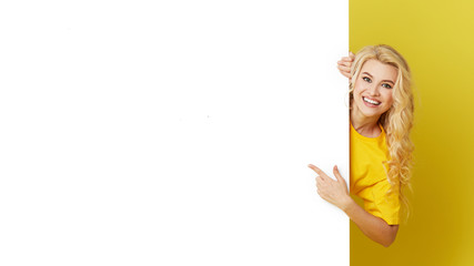 Wall Mural - Young happy woman peeks out from behind a white banner on a yellow background. Point to an empty blank on a form, a copy space for text. Horizontal shot