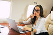 © My Ocean studio - A nice businesswoman in formal attire and glasses, who is typing something on her laptop with thoughtful face expression, touching her chin while flying on a private jet.