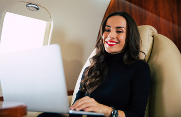 Radiant smile. Close-up photo of a gorgeous girl with perfect brown hair, who is smiling, while typing something on her laptop during the flight.