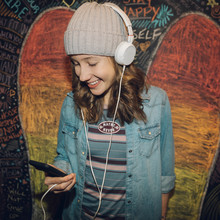 Smiling Caucasian Tween Girl With Headphones And Smart Phone Listening To Music Against Wall With Chalk Wings