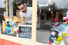 Male Business Owner Placing Help Wanted Sign In Art Supply Shop Window