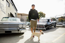 Portrait Latinx Young Man With French Bulldog In Parking Lot With Low Rider Cars